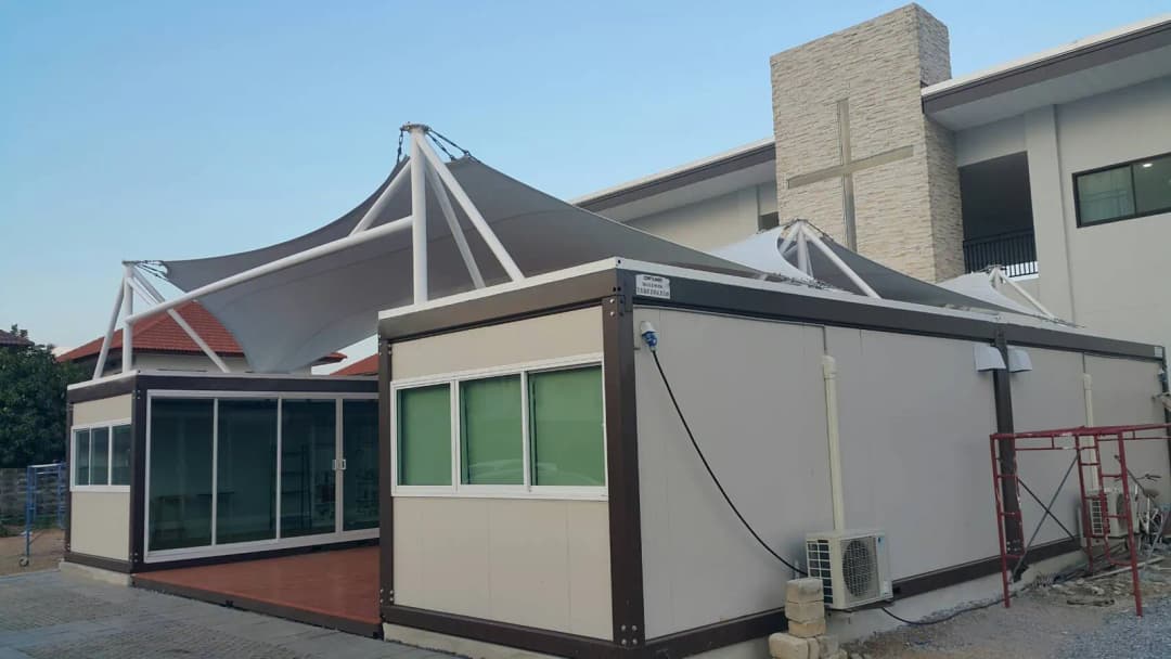 ノックダウンの建物。クリームと茶色のユニット2列、中央にウッドデッキ、白い膜構造の屋根、教会の前(3枚目)