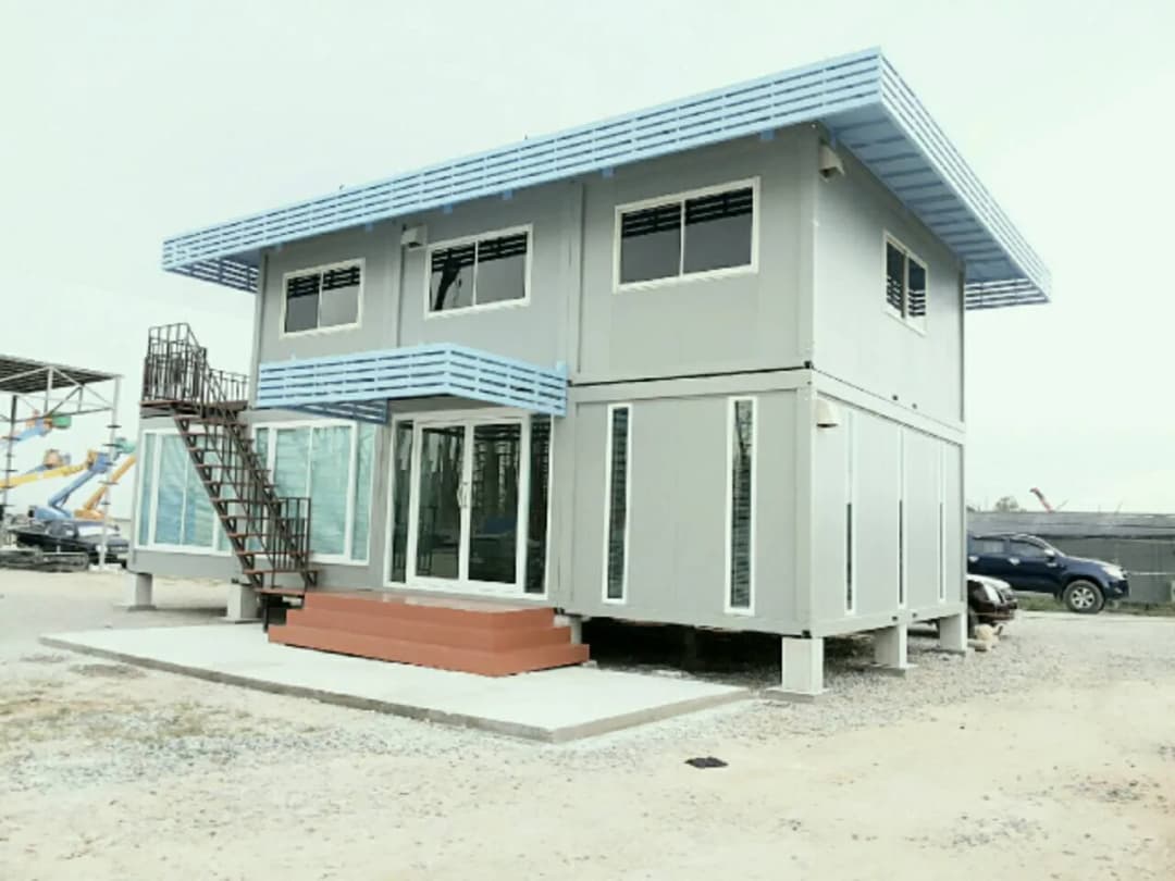 グレーの2階建てノックダウンハウス。水色のルーバー、1階に張り出したガラスの部屋、鉄骨階段(3枚目)