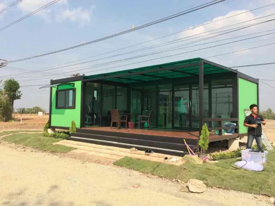 ノックダウンのカフェ。緑の壁、全面ガラス、ポリカーボネート屋根、ウッドデッキ、テーブルと椅子