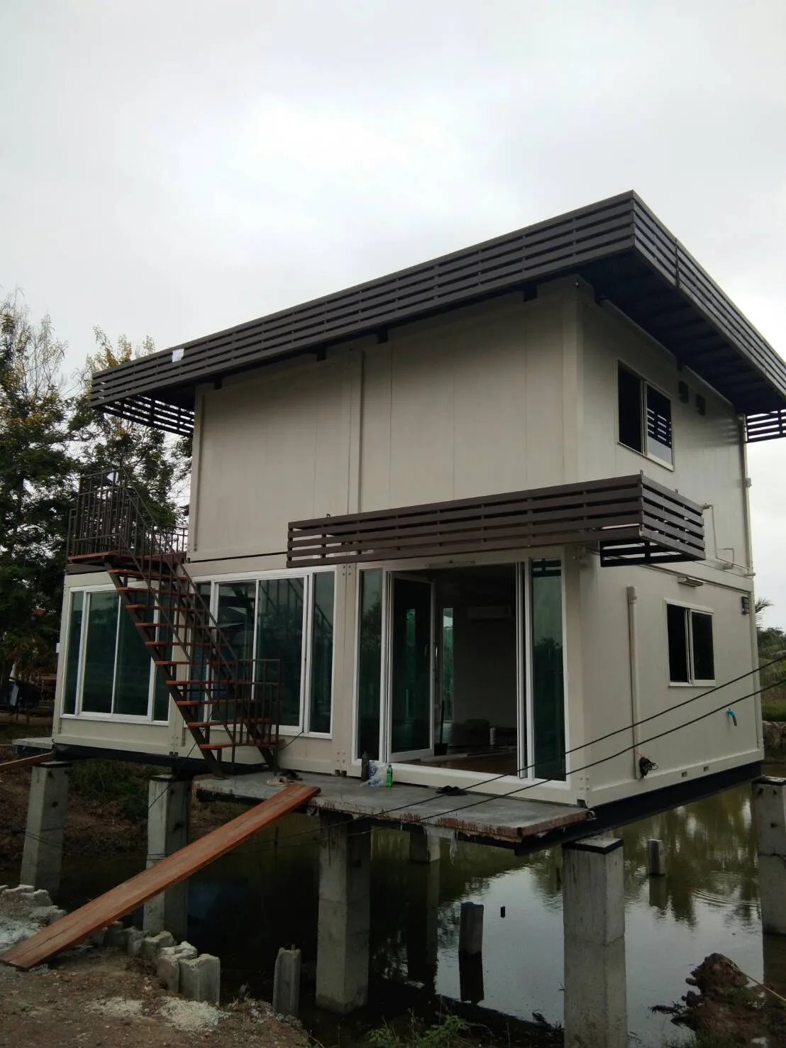 Two-storey knock-down house on concrete piles over a pond, glass ground floor, steel staircase