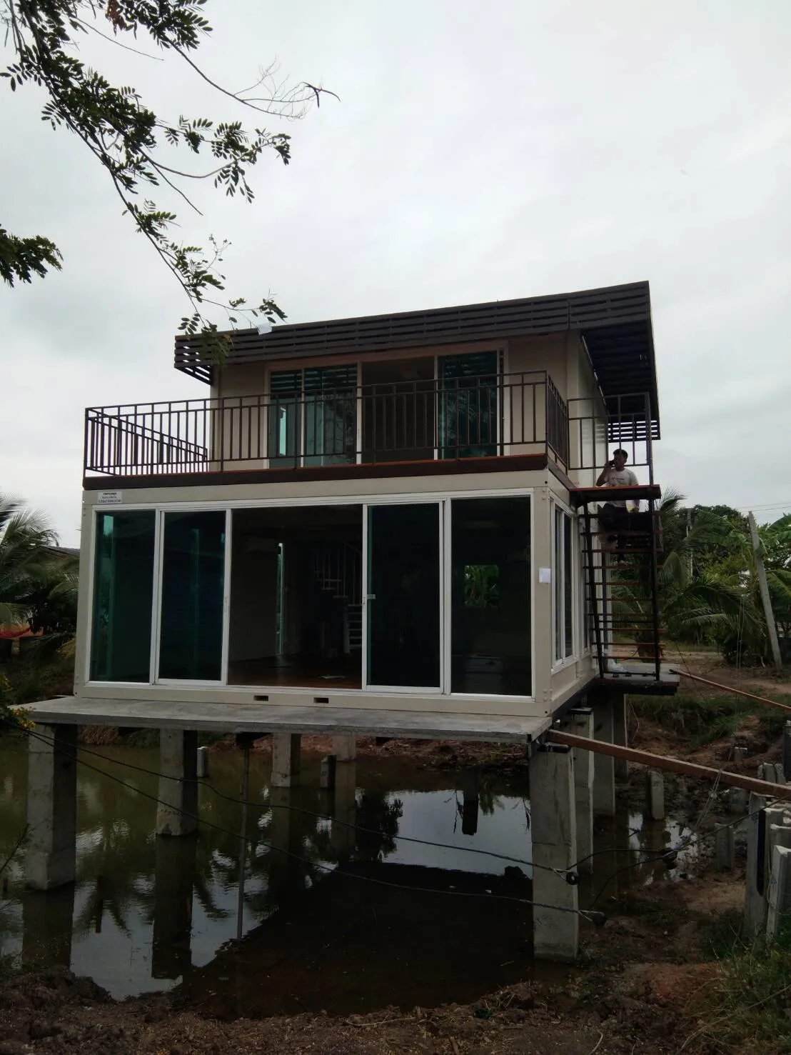 Two-storey knock-down house on concrete piles over a pond, glass ground floor, steel staircase (view 2)