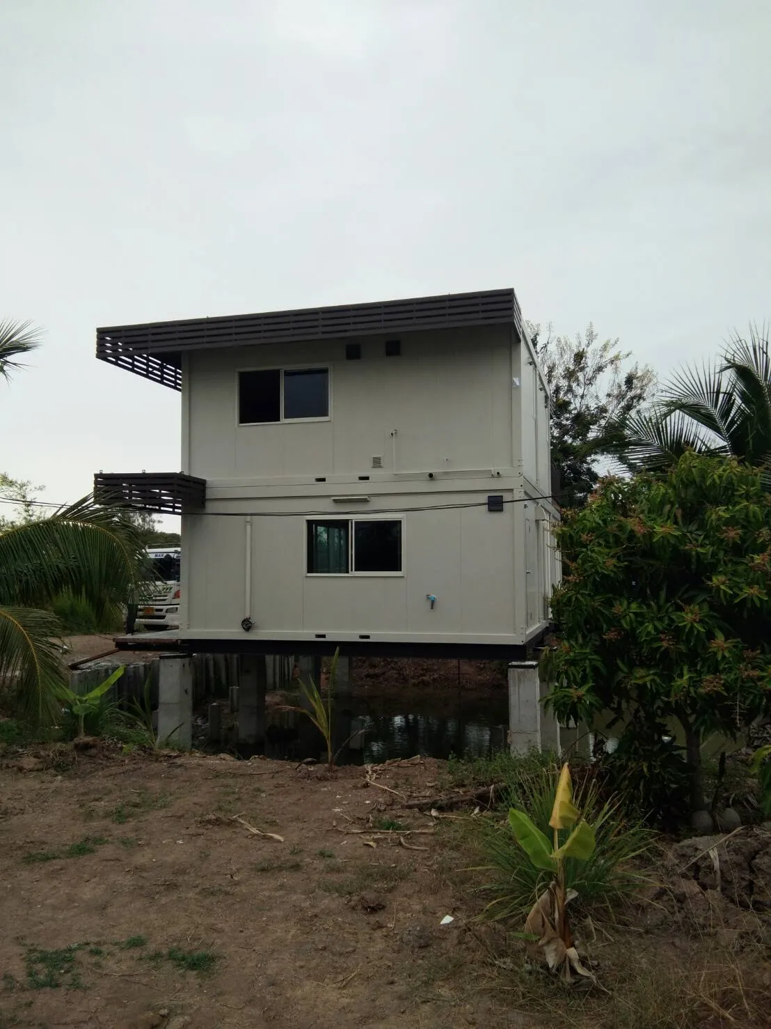 Two-storey knock-down house on concrete piles over a pond, glass ground floor, steel staircase (view 3)