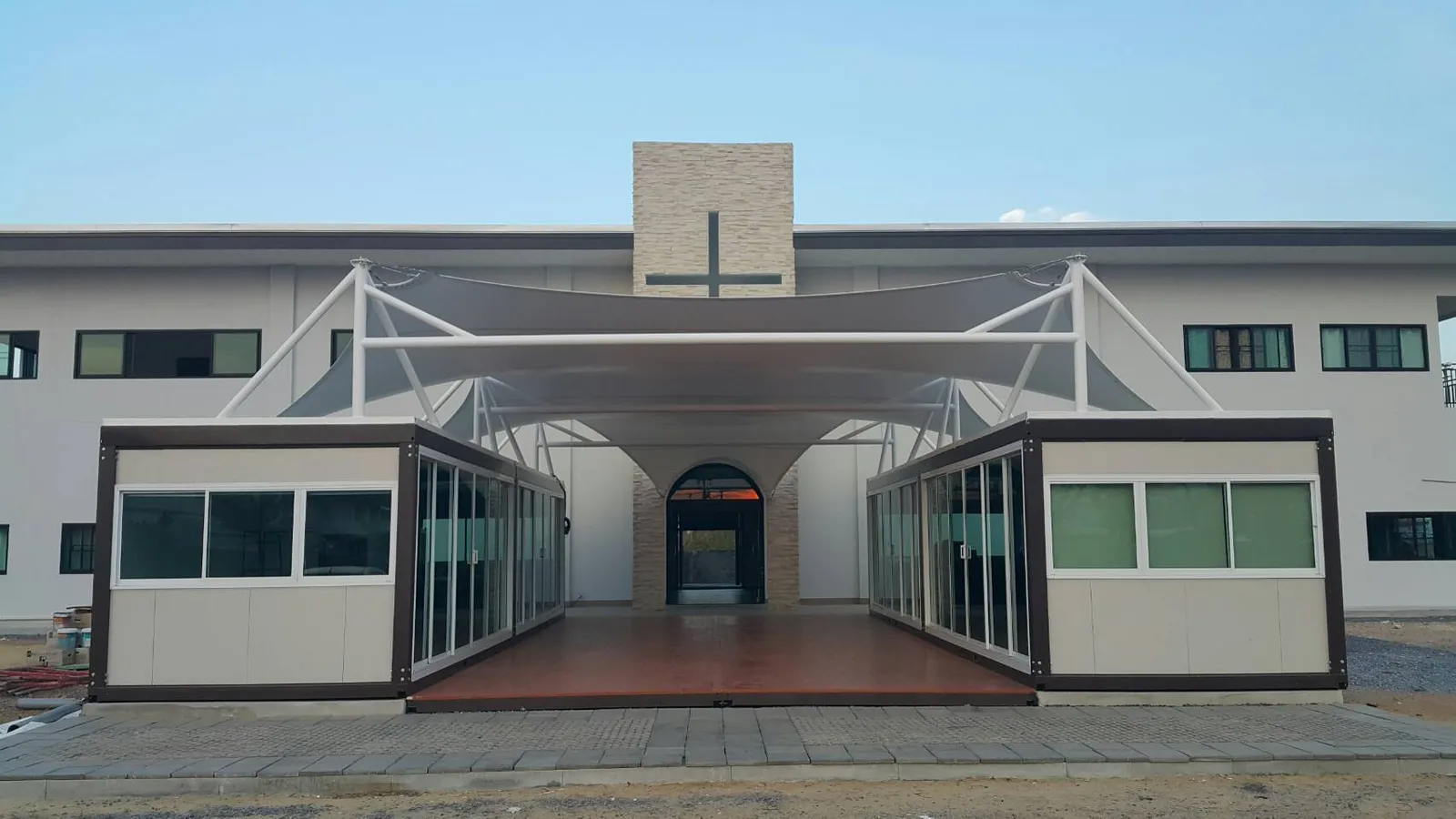 Knock-down hall: two rows of cream units with a wooden deck between and a white tensile-fabric roof, in front of a church