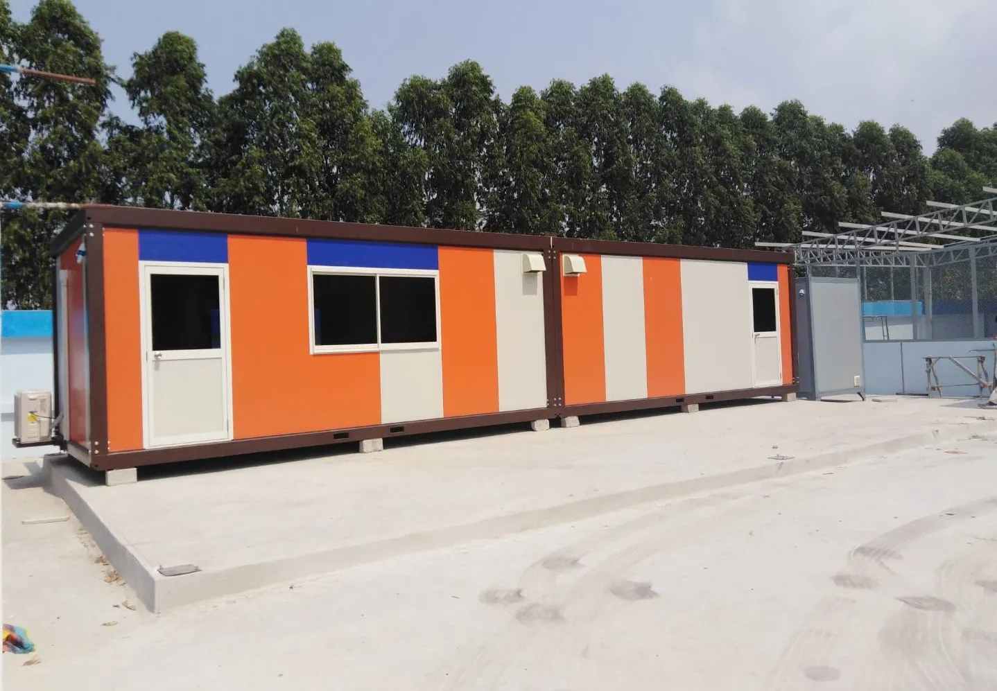 Knock-down worker housing joined in a long row with orange, white and blue striped walls on a concrete yard (view 2)