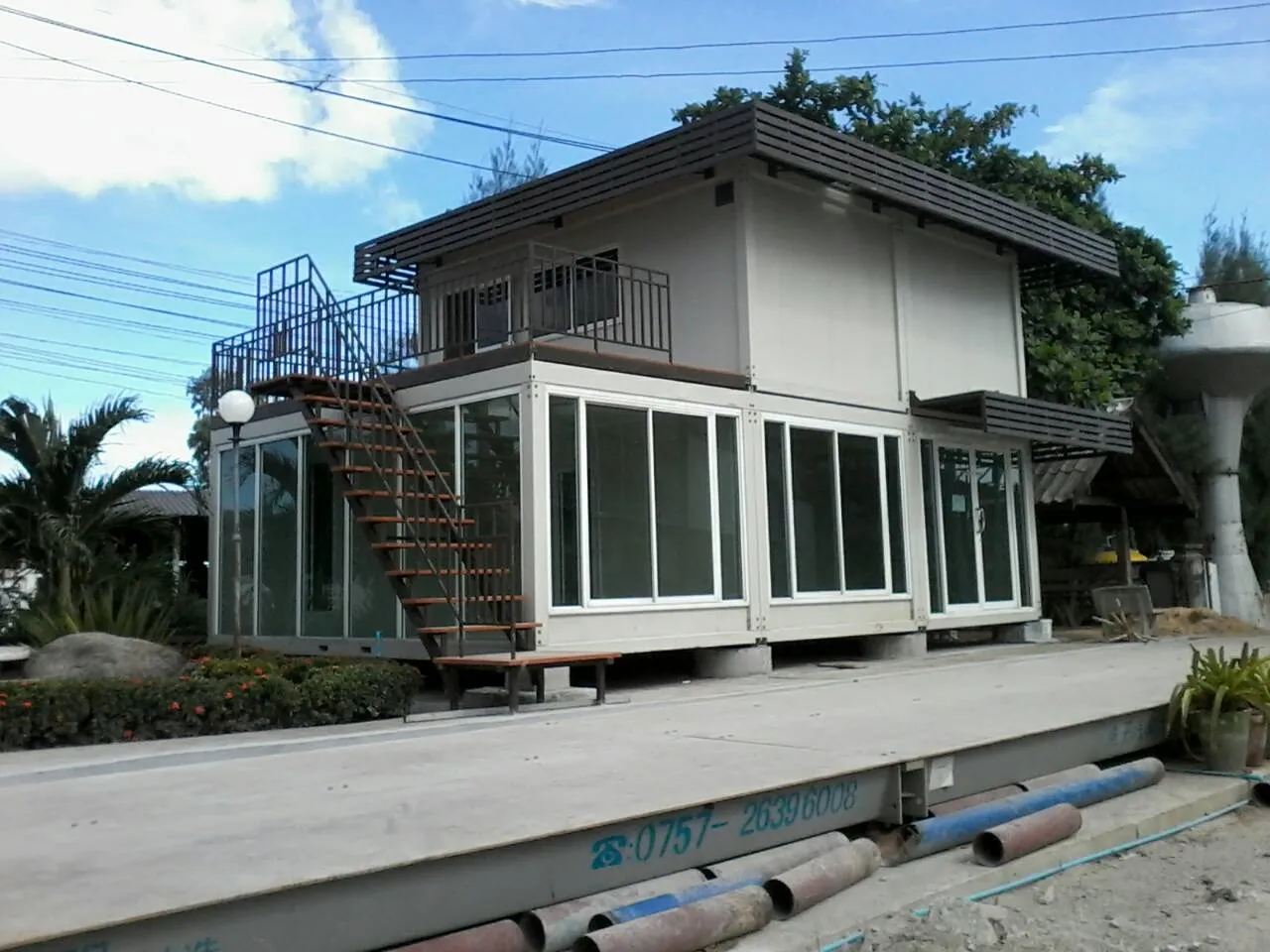Two-storey knock-down house in white and dark brown with sun louvres and an upper balcony, set in a garden